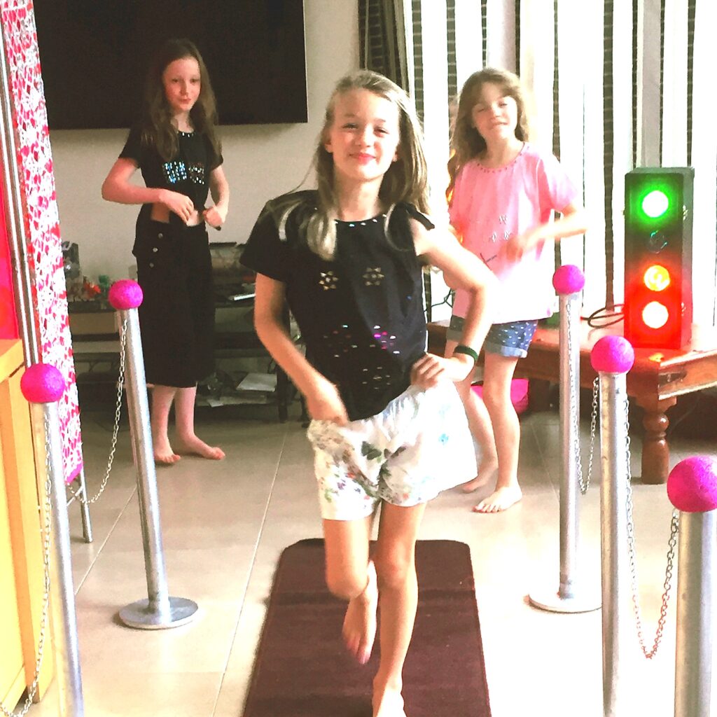 Three girls, one is strutting down a carpet runner like a model whilst two friends are in the background.