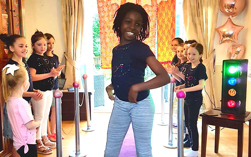 Girl posing for a photograph with a large smile on her face. disco lights and girls are in the background clapping.