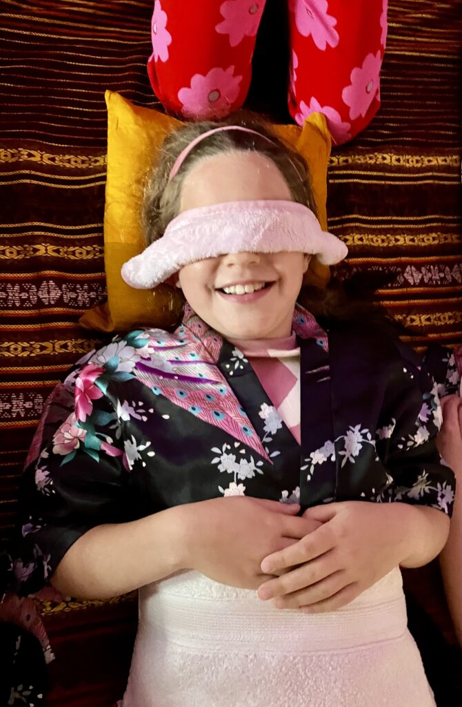 Girl smiling to camera with a pink eye mask on. She is lying down with oriental gown on with her head on a yellow pillow.