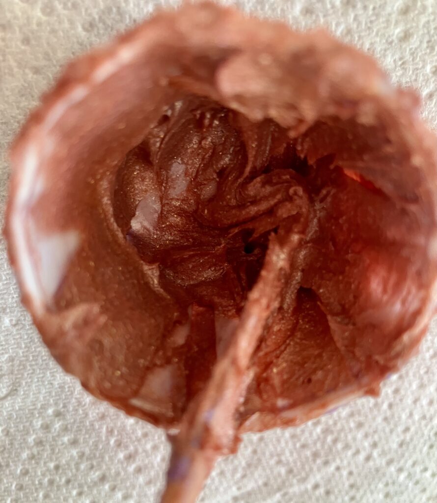 Rose gold coloured lip gloss in a pot with a stirring stick.