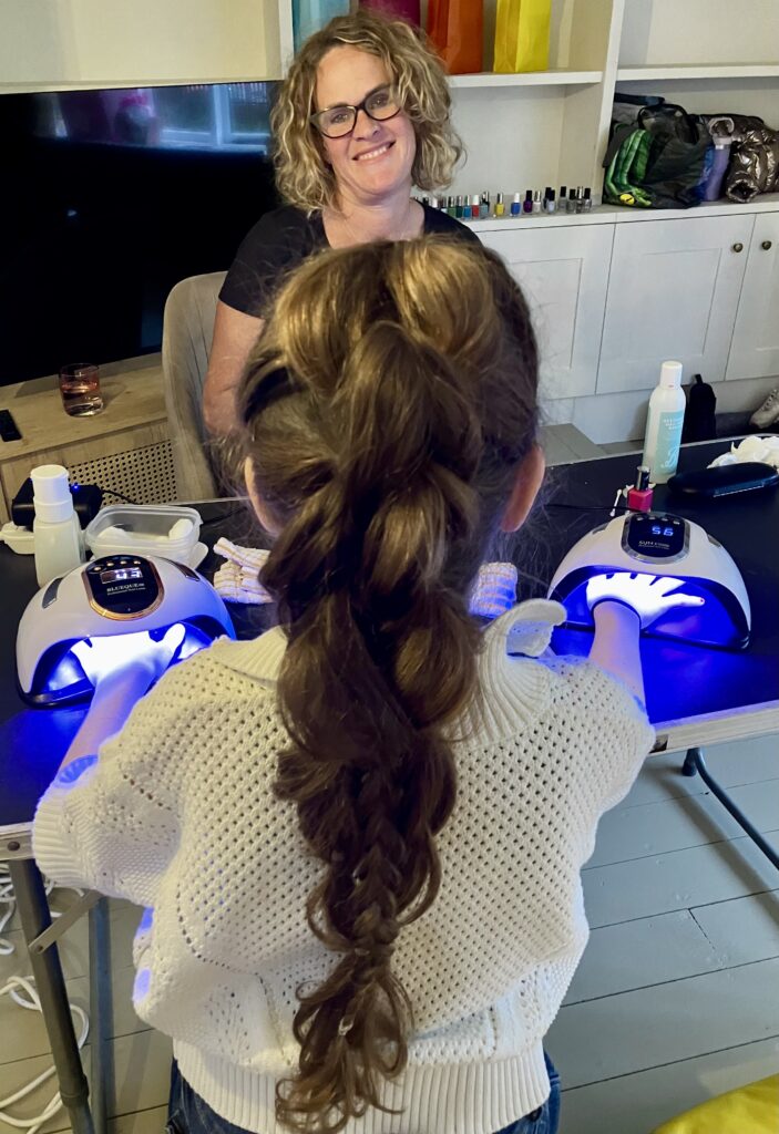 Girl having her nails painted at a table with a giddy diva staff sat opposite smiling at her.