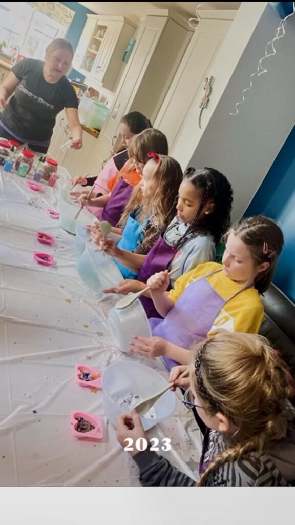 Six girls lined up along the table with bowls making bath-bomb.