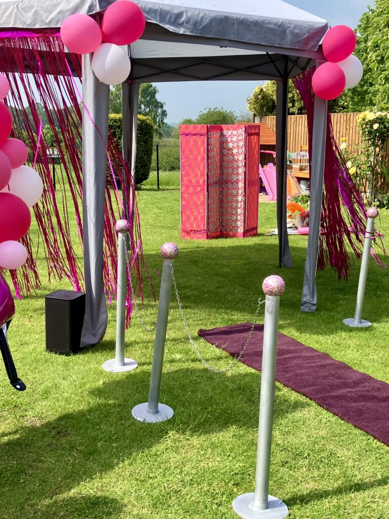Maroon runway and silver chrome poles with pink glittery balls line either side of the catwalk runway. It's a sunny day in a garden with a silver gazebo with balloons & a pink transformation screen in the background.