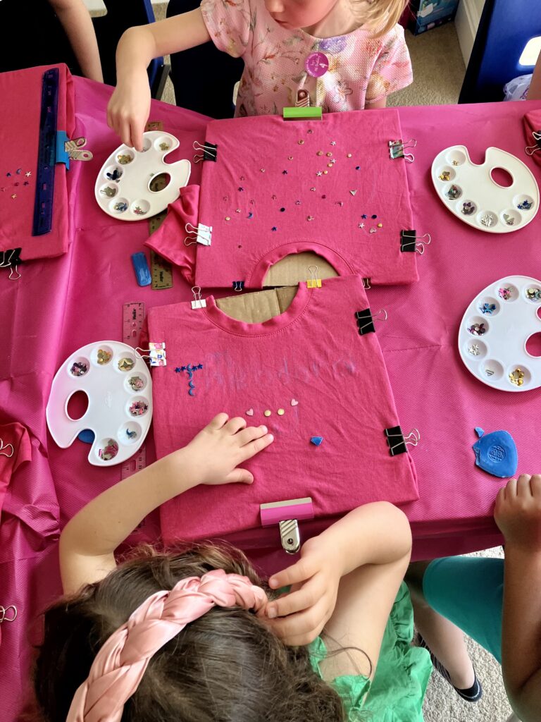 Two girls sat opposite each other at a table placing rhinestone gems on pink t-shirts for decoration