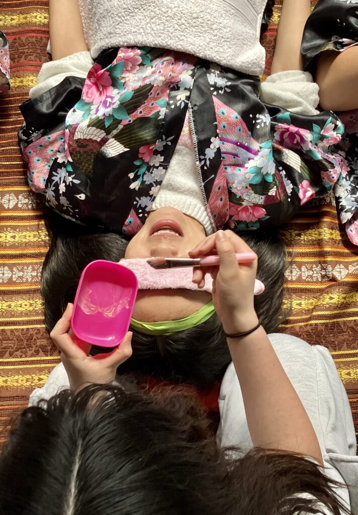 Girl lying down wearing an oriental gown and a pink eye mask. She is having a face mask applied to her face with a brush.